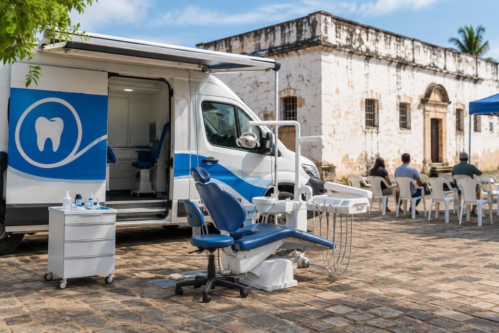 Van odontológica branca e azul estacionada em área de chão de pedra com cadeira odontológica ao ar livre, em frente a prédio antigo com paredes brancas. Pessoas sentadas em cadeiras plásticas brancas aguardam atendimento.