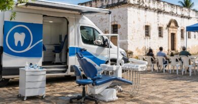 Van odontológica branca e azul estacionada em área de chão de pedra com cadeira odontológica ao ar livre, em frente a prédio antigo com paredes brancas. Pessoas sentadas em cadeiras plásticas brancas aguardam atendimento.