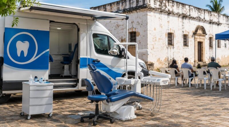 Van odontológica branca e azul estacionada em área de chão de pedra com cadeira odontológica ao ar livre, em frente a prédio antigo com paredes brancas. Pessoas sentadas em cadeiras plásticas brancas aguardam atendimento.