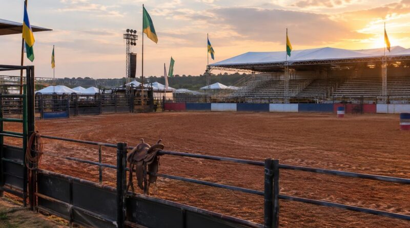 Arena de rodeio ao ar livre com chão de terra batida, cercada por grades e com arquibancadas ao fundo. Bandeiras verdes, amarelas e azuis estão fincadas em postes ao redor da arena. O céu ao fundo mostra a luz do pôr do sol, criando atmosfera calma e acolhedora.